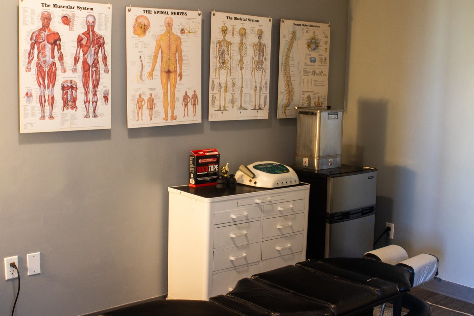 Chiropractic treatment room with an adjustment table, e-stim and ultrasound units, and anatomy charts